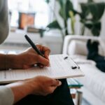 Unrecognizable ethnic female therapist taking notes on clipboard while filling out form during psychological appointment with anonymous client lying on blurred background